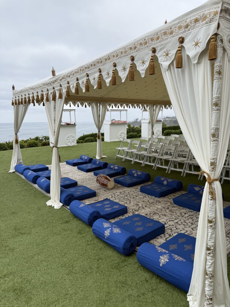 White and gold event tent with blue floor cushions, decorative tassels, and oceanfront lounge seating.