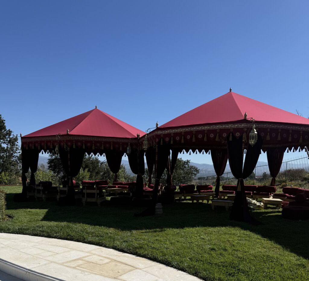 Red tent canopies with decorative trim and hanging black curtains set up on a grassy lawn under a clear blue sky, with mountains in the distance.