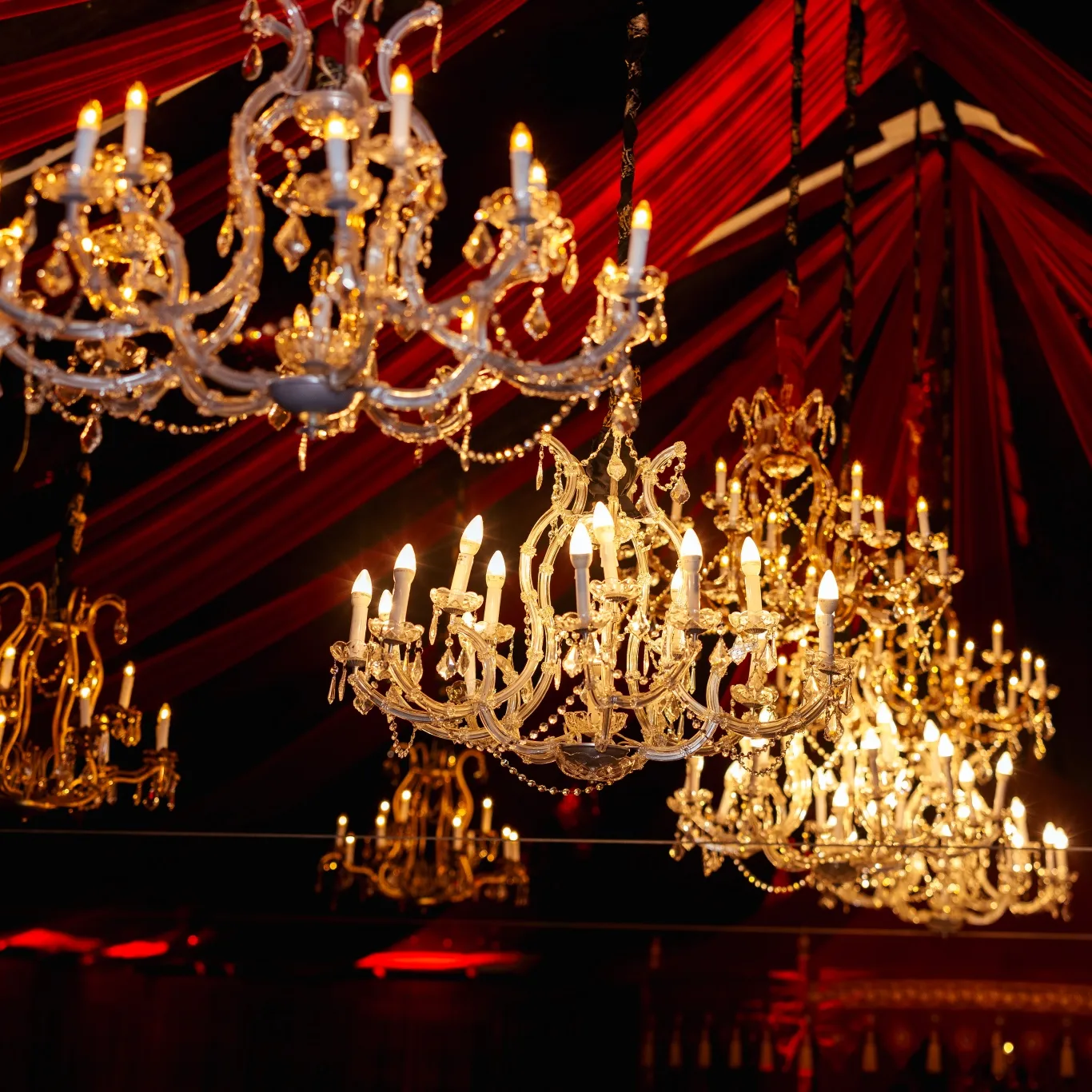 Crystal chandeliers hanging from a red-draped ceiling, lit by warm candle-like bulbs at a luxurious event venue.